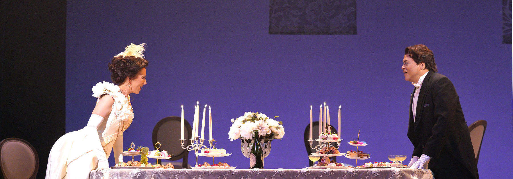image from a performance of a couple sitting at opposite ends of a dining table